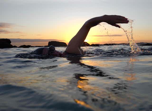 A silhouetted swimmer raising their arm out of the water backlit by a sunset