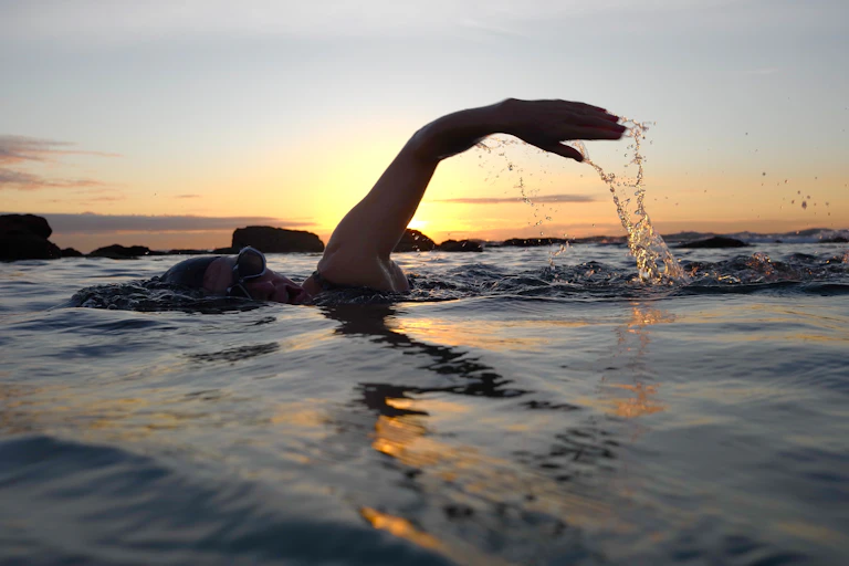 A silhouetted swimmer raising their arm out of the water backlit by a sunset
