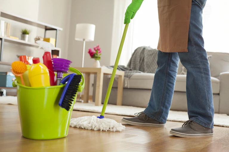 Floor-level view of a person's legs and a bucket of cleaning equipment in a sitting room. The person is wearing an apron and gloves, and pushing a mop.
