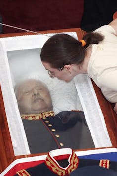 Augusto Pinochet yace muerto en el ataúd con tapa transparente, una chica se acerca.
