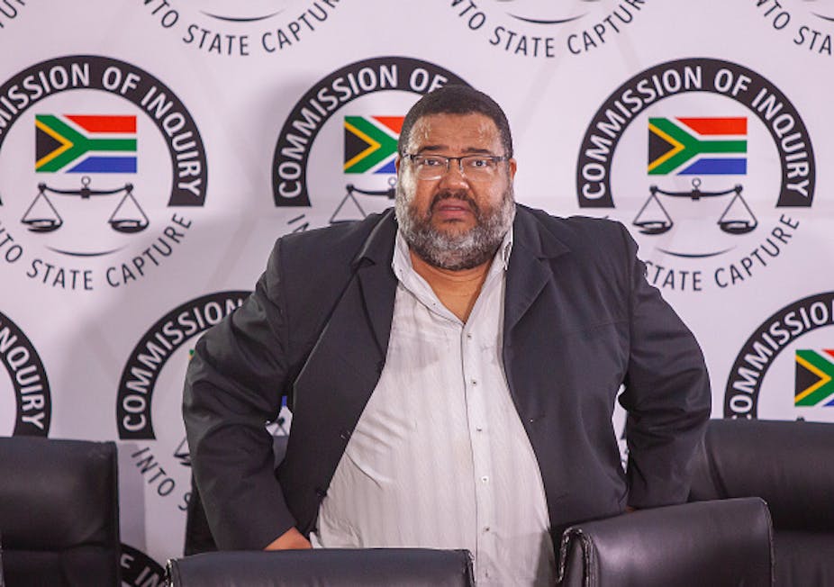A man wearing a jacket and shirt without a tie is photographed standing in front of the State Capture Commission emblem. 