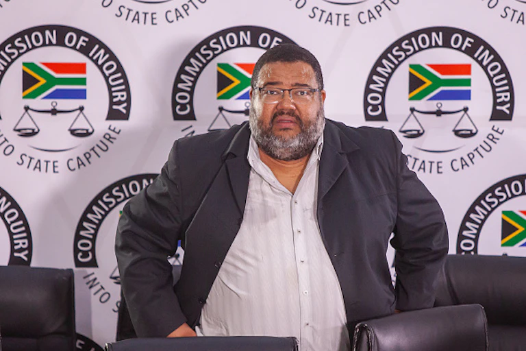 A man wearing a jacket and shirt without a tie is photographed standing in front of the State Capture Commission emblem.