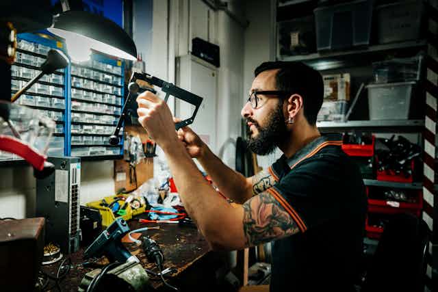 A young man with glasses, dark hair, a beard and arm tattoos looks at parts while working in a workshop.