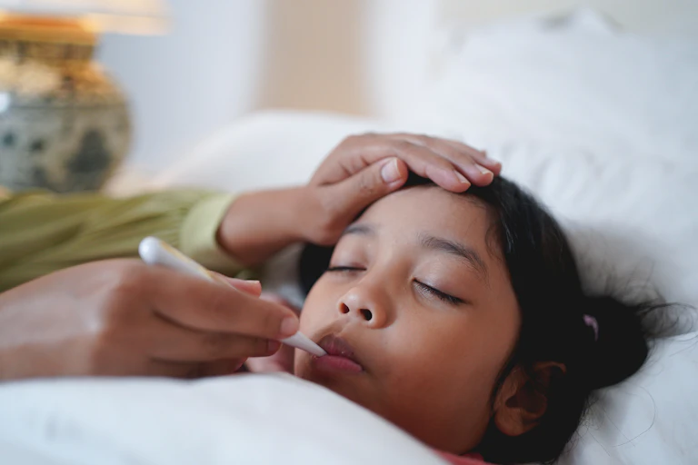 Sick child tucked under the covers, with someone taking their temperature with a thermometer.