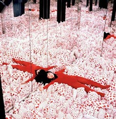 Una mujer vestida de rojo tumbada sobre globos blancos con topos rojos en una habitación llena de espejos.