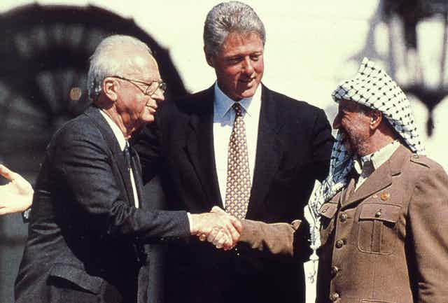 A man in traditional Palestinian headscarf shakes hand with a suited man in glasses while a tall man with gray hair stands between