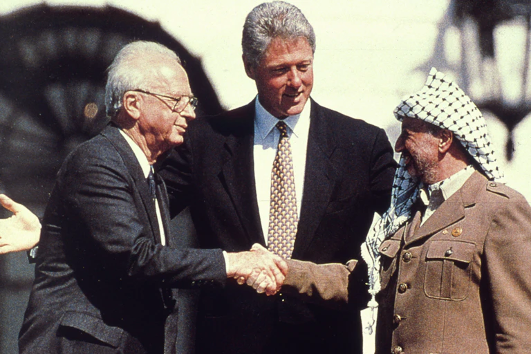 A man in traditional Palestinian headscarf shakes hand with a suited man in glasses while a tall man with gray hair stands between.