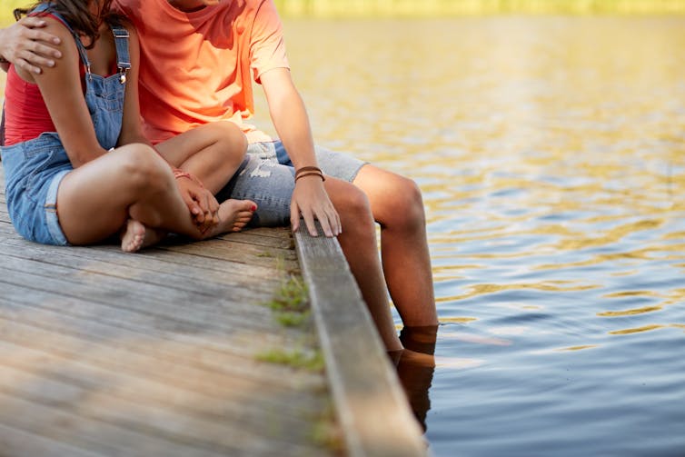 teen couple sits together on pier near water