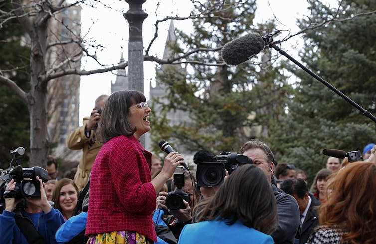A woman in a red jacket smiles while speaking into a microphone as people snap photos of her outside.