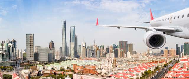An airplane is shown flying over low-rise residential buildings, with the skyscrapers of Shanghai behind.