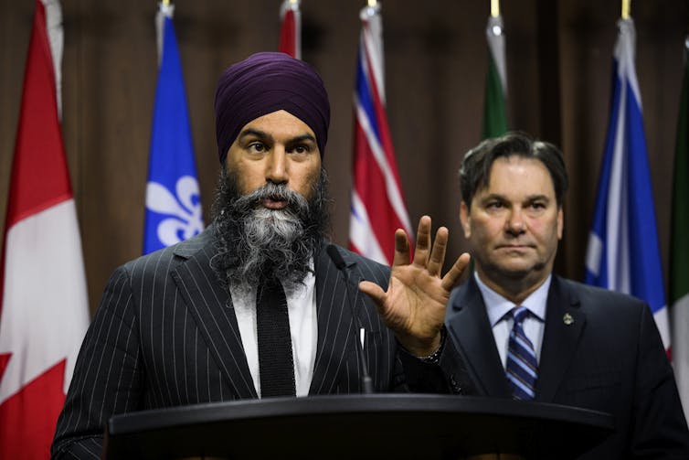 Jagmeet Singh speaking behind a lectern with health critic Don Davies beside him, in front of a row of flags