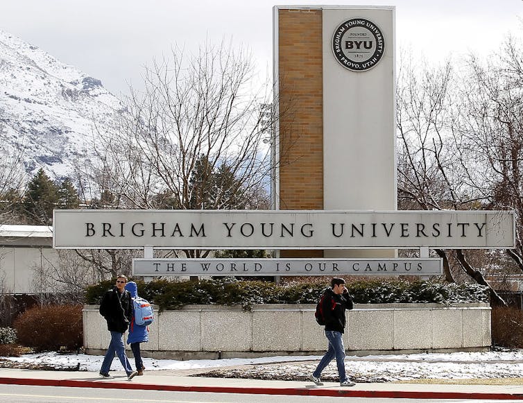 A few students walk by a sign for Brigham Young University with snowy mountains in the distance.
