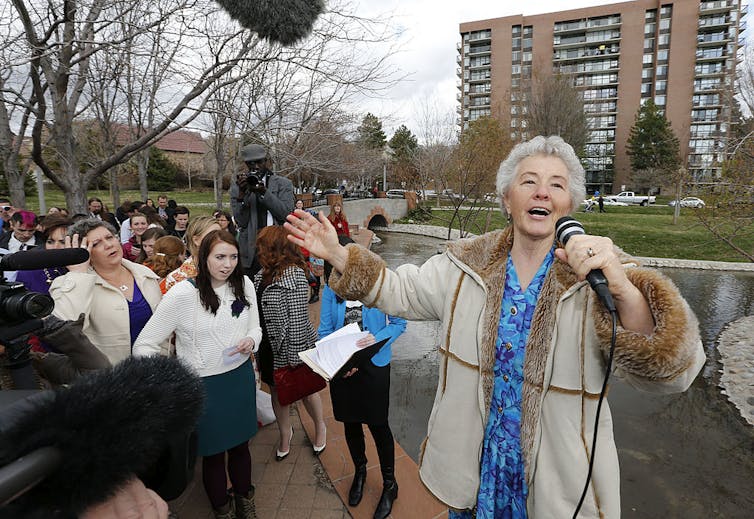 An older woman in a coat and blue dress sings into a microphone on an overcast day, with a small group of women visible behind her.