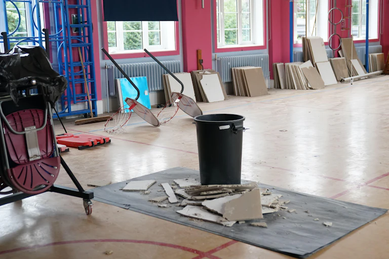 A large, empty room in a school with chunks of Raac ceiling that have collapsed onto the floor