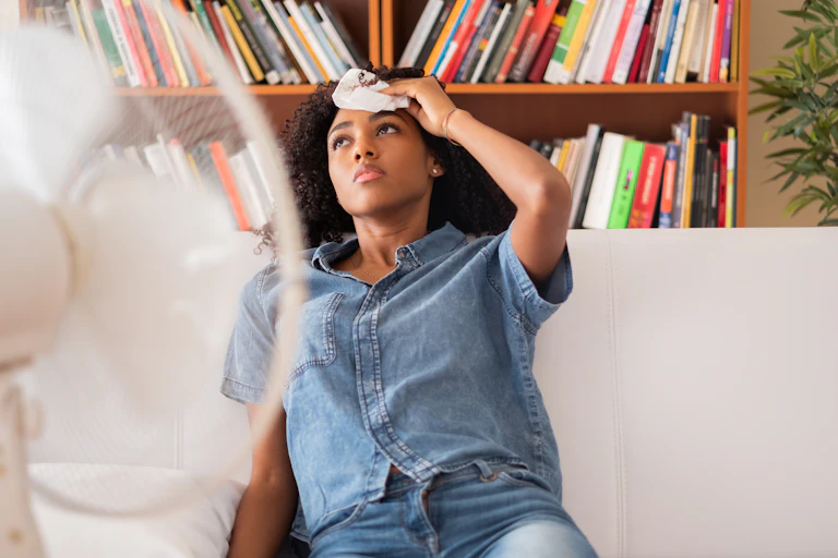 A woman struggling with the heat sits on a couch in front of a fan with a cloth on her head.