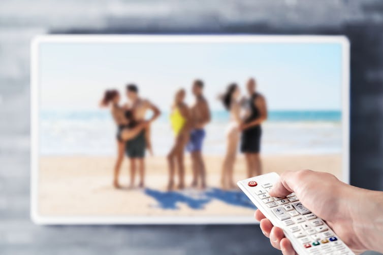 Blurred image of three couples on a beach on a tv screen, with hand holding remote control in the foreground.