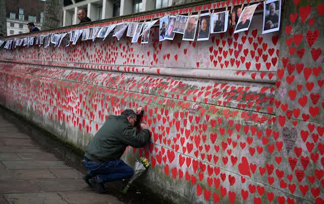 A man leaving flowers laying his hands and head on a wall covered in red hearts to commemorate every British person who died in the pandemic.