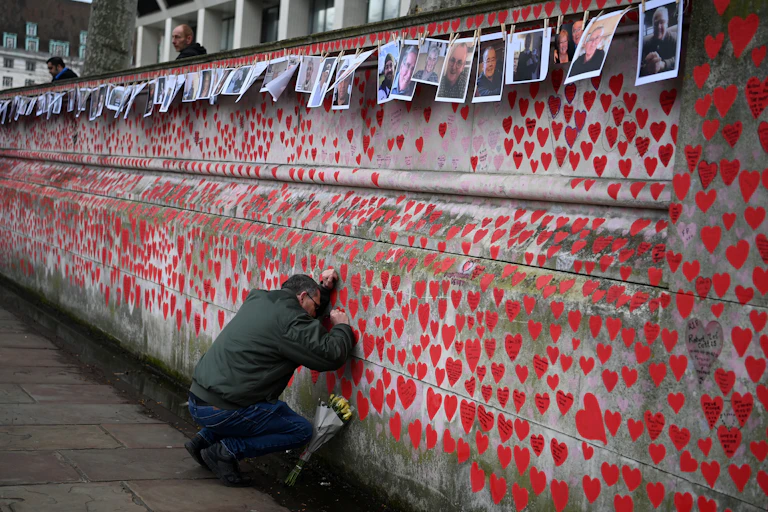 A man leaving flowers laying his hands and head on a wall covered in red hearts to commemorate every British person who died in the pandemic.