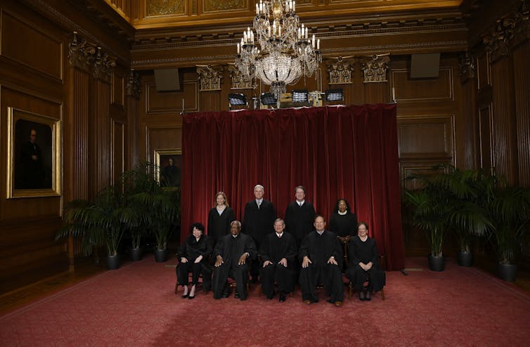 Nine people in black robes seated together in an elegant, high-ceilinged room under a chandelier.