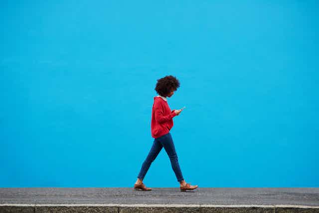 A young woman in a red jersey and an Afro hairstyle walks past a blue wall looking at her cellphone.