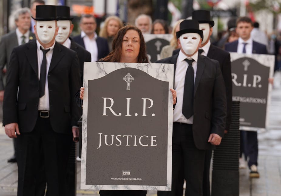 Protesters dressed in funeral wear and white masks are led by a woman holding an image of a headstone reading 'RIP Justice'.