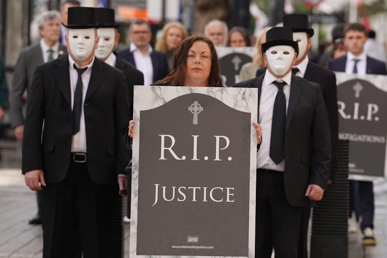 Protesters dressed in funeral wear and white masks are led by a woman holding an image of a headstone reading 'RIP Justice'.