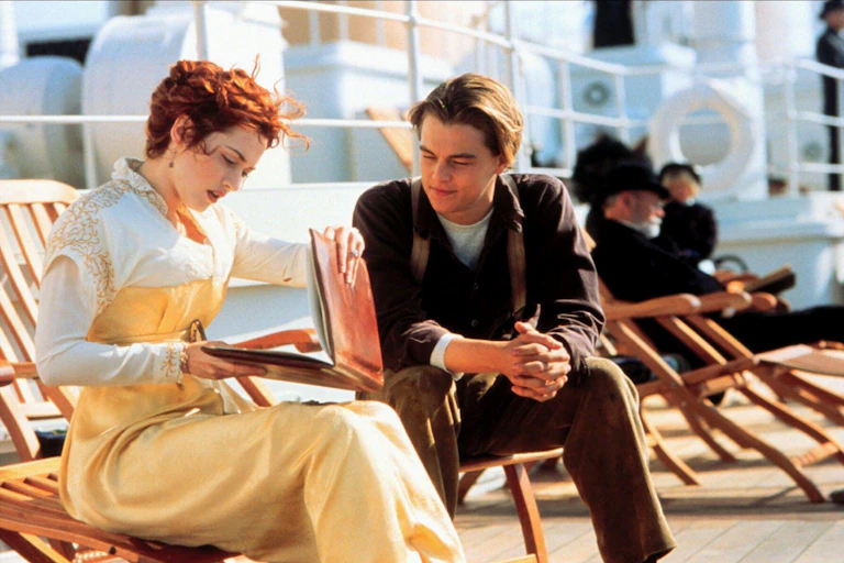 Film still of Kate Winslett (Rose) and Leonardo DiCaprio (Jack) on the Titanic, characters are sitting in deckchairs looking at a notebook.