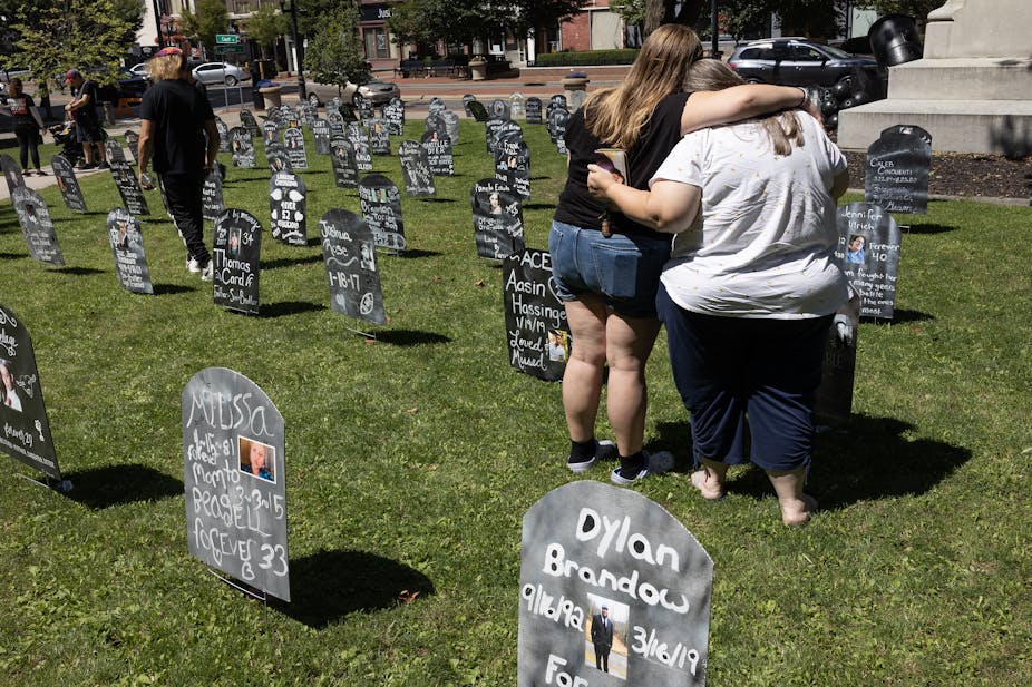 Two women with their backs to the camera hug as they view a replica tombstone and other people walk by