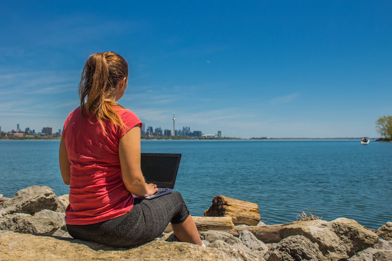 A woman working on a laptop sits by a lake. The Toronto skyline is in the distance.