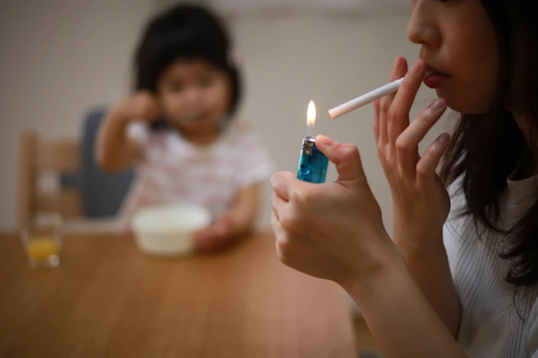 Uma mãe acende um cigarro perto de sua criança, que está sentada e comendo ao fundo