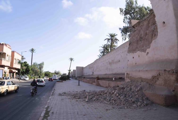 A brick ancient wall with rubble scattered near it and some intact buildings across.