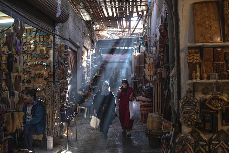 Shops selling an array of colorful goods on either side of a narrow old marketplace while two women in headdress walk through the lane in the center.