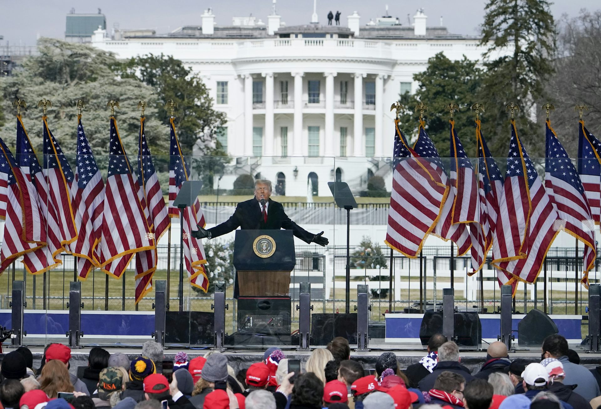 A man in front of a presidential podium gestures as he speaks. A large white mansion is behind him.