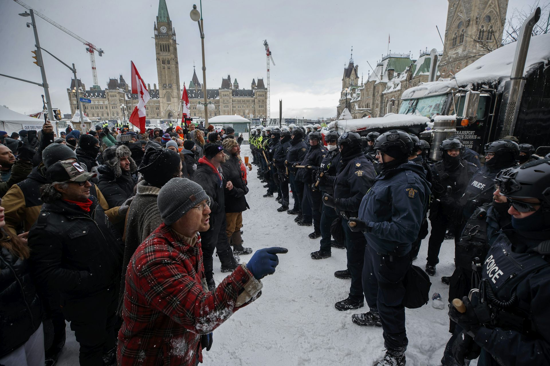 A row of protesters stand in front of a row of armoured police officers. One man points his finger at police.