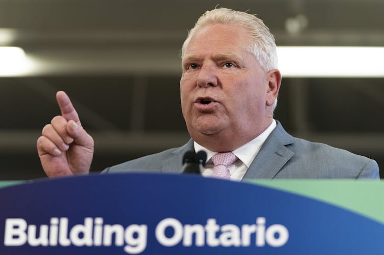 A man with slicked-back white hair points while speaking behind a banner that reads Building Ontario.