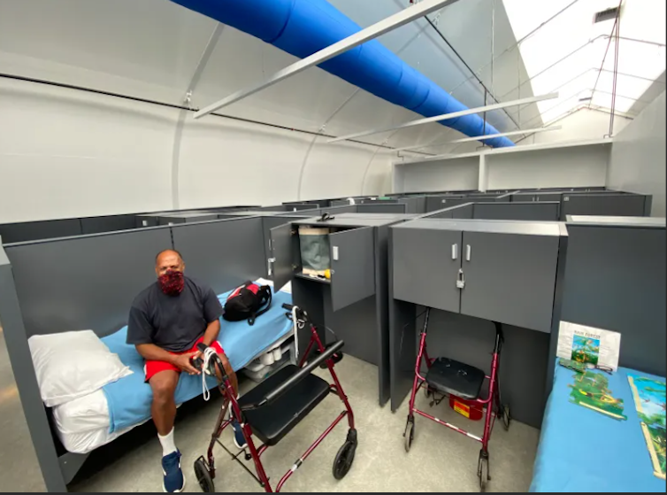 A man sits on a bed in a large room divided into individual spaces with low partitions. His area has storage compartments with locks and a skylight provides daylight.