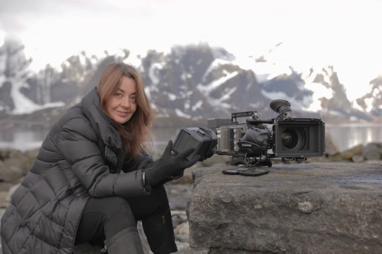 A woman wearing a heavy dark blue jacket and gloves is pictured against the backdrop of snow-covered mountains. She is holding part of a film camera.