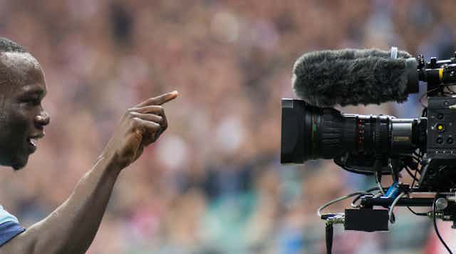 A man, sweating from exertion in a stadium full of people points a finger directly at a television camera