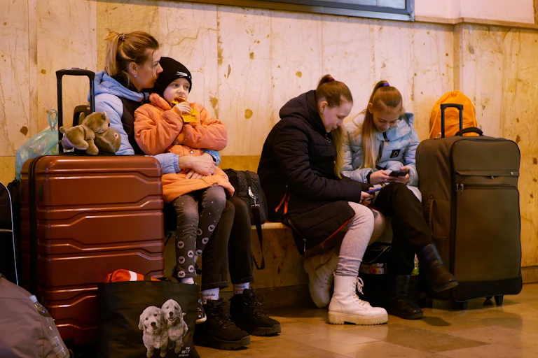 A mother holds a young child on her lap surrounded by bags. Two young girls sit next to them, also with bags.