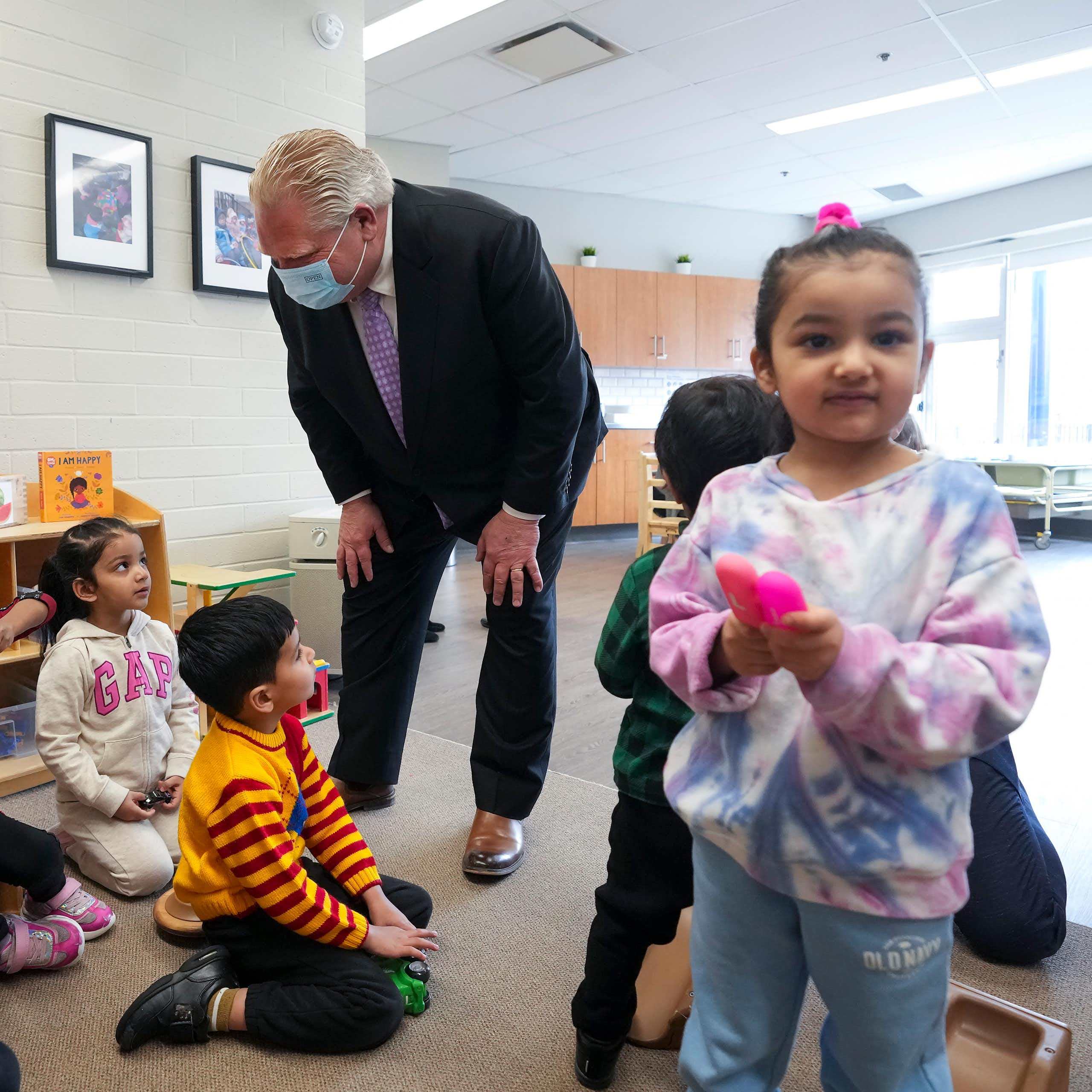 A man in suit and face mask seen bending over talking with children as a child holds a pink object in the foreground.
