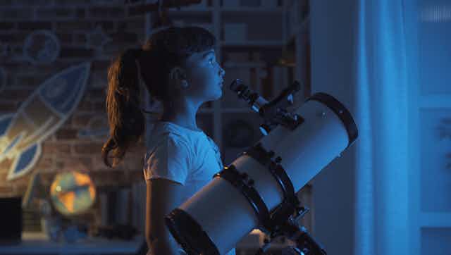 Seorang gadis berdiri di dekat teleskop dan melihat ke luar jendela pada malam hari