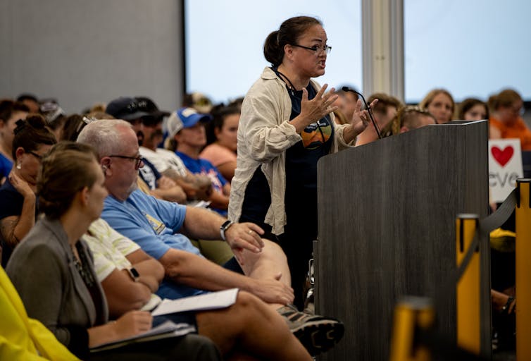 A person standing at a lectern gestures and makes an emotional facial expression.