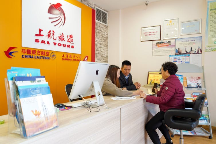 Beneath a sign reading Sal Tours, a man and woman behind a desk show paperwork to a woman in front of the desk.