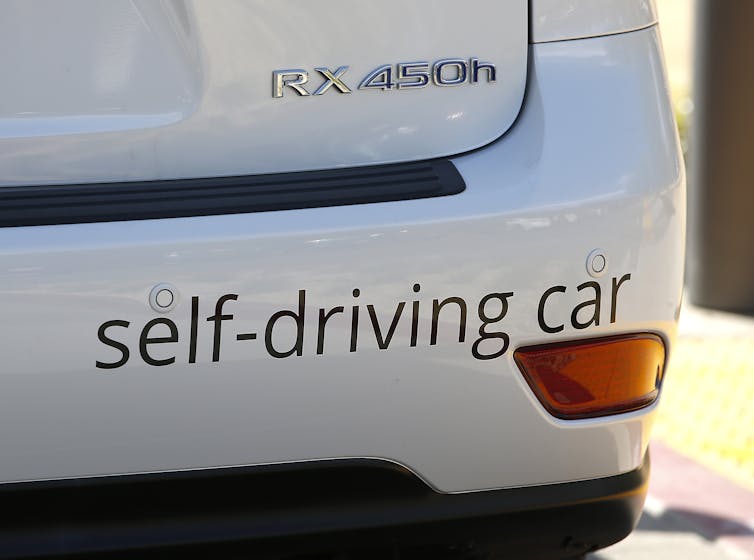 A zoomed in shot of a white car with a bumper sticker reading 'self-driving car'