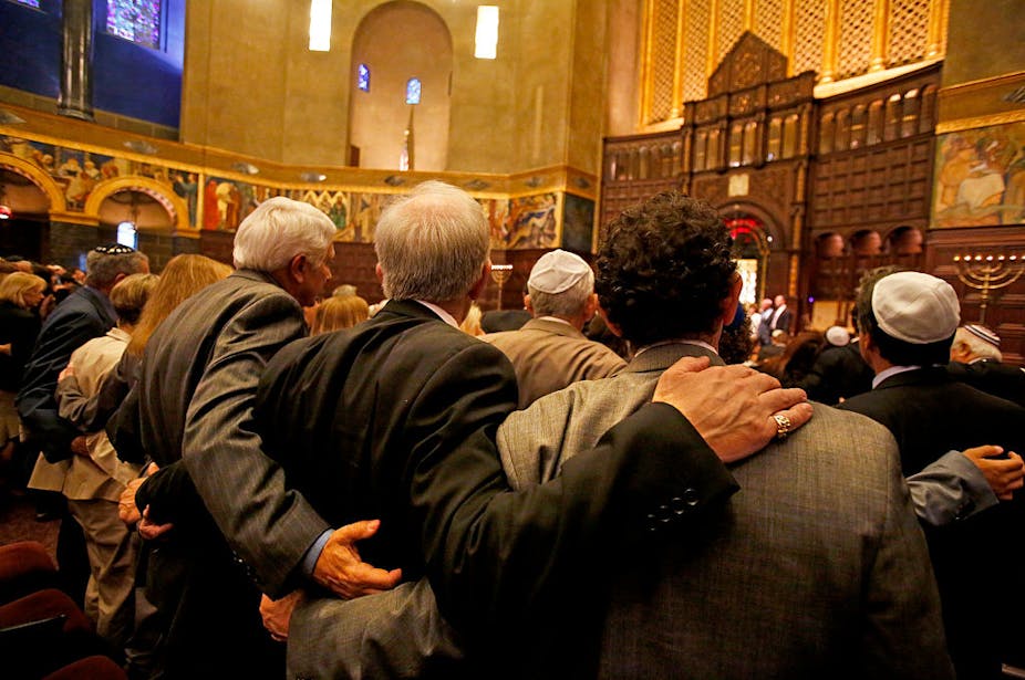 A packed congregation, seen from the back, as men stand with their arms around each other.