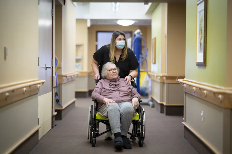 A woman in a medical mask pushes an older woman down a hallway in a wheelchair