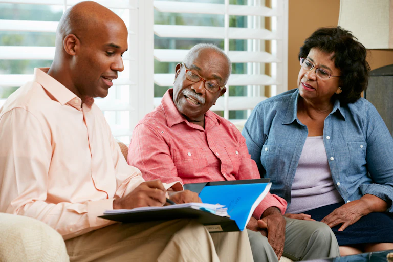An elderly couple look at an open document on a young man's lap. The young man is speaking and pointing at the document with a pen