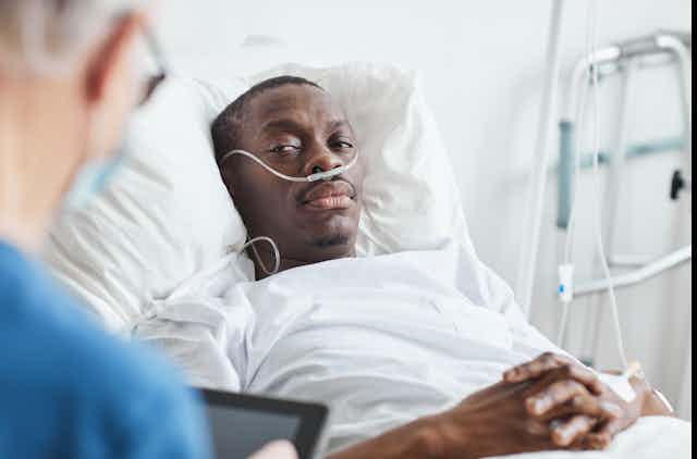 A Black man wearing a nasal cannula rests in his hospital bed while his doctor talks to him.