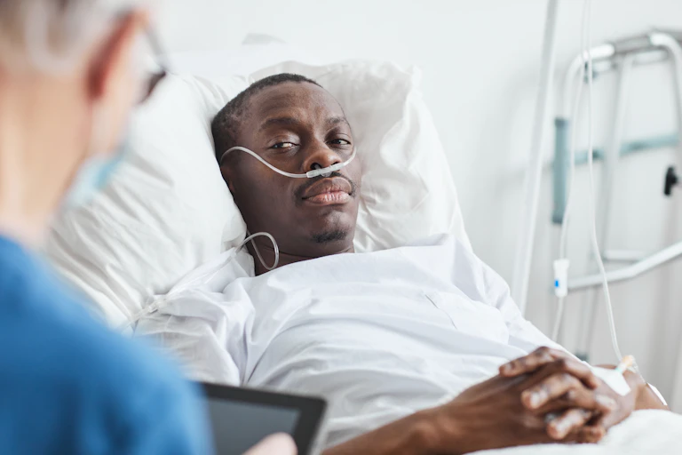 A Black man wearing a nasal cannula rests in his hospital bed while his doctor talks to him.