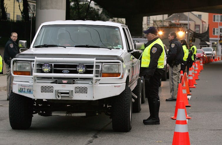 A long line of uniformed police officers stand with their backs to range cones as they speak individually to the motorists in the cars parked in front of them.